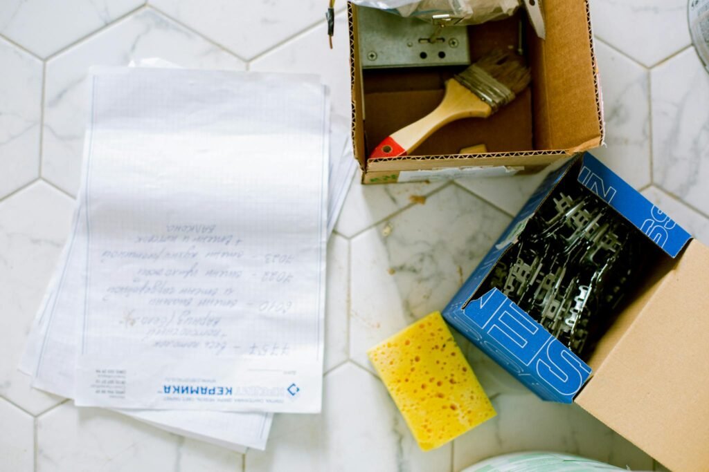 Flat lay of home renovation tools including paintbrush, sponge, and hardware on tiled floor.